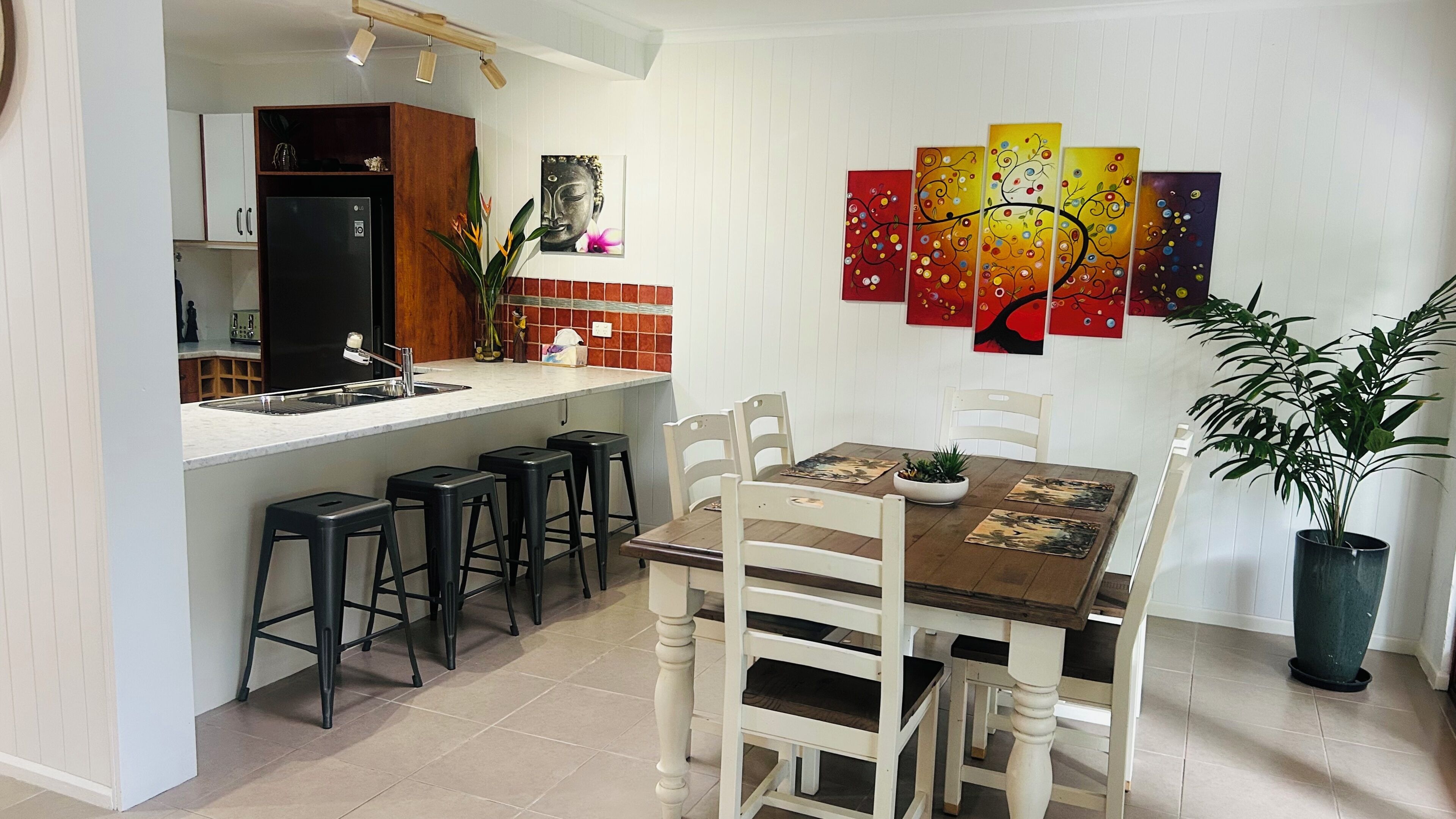 A modern kitchen with a built-in refrigerator, bar stools, a dining table with chairs, and a potted plant.