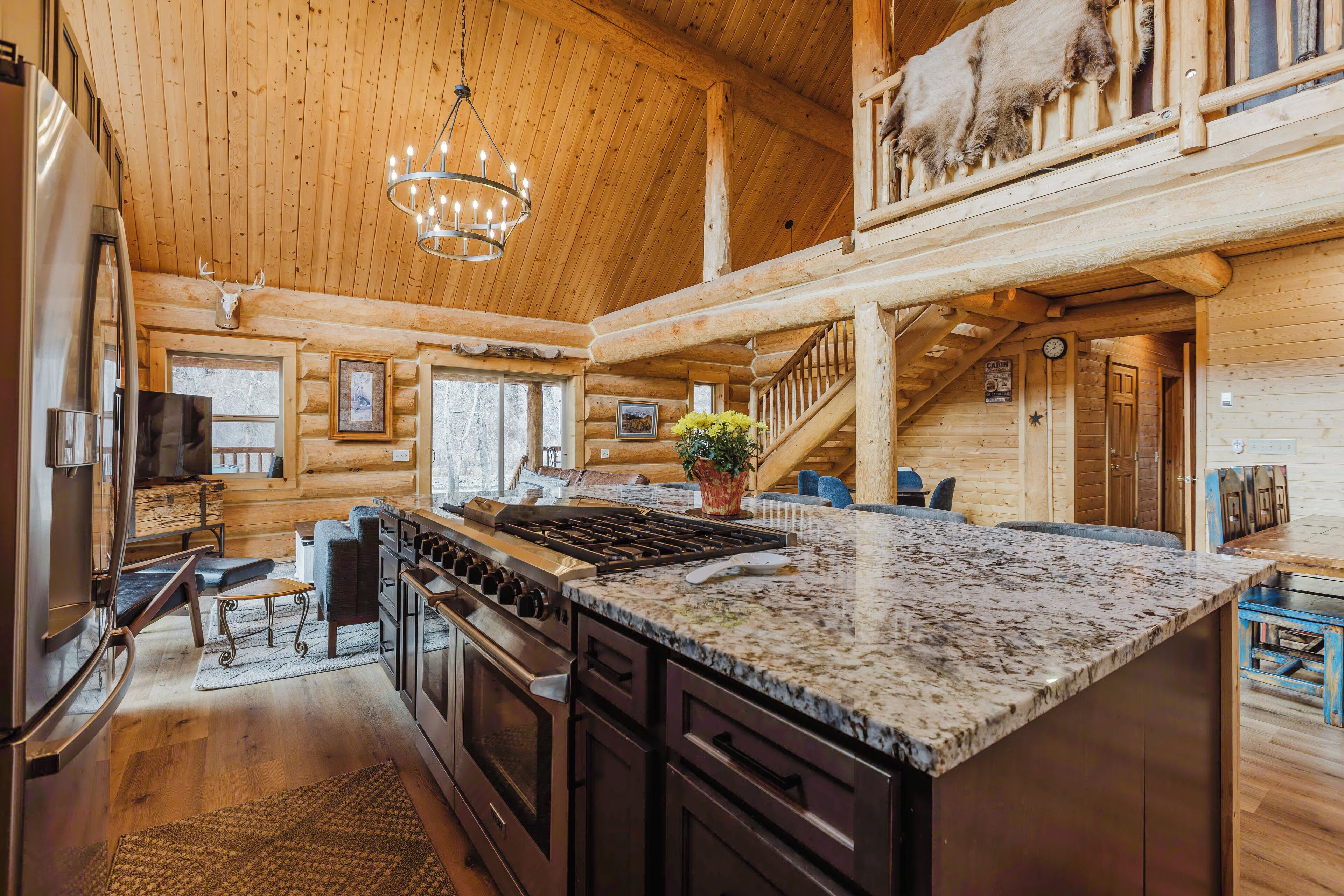 Une cuisine spacieuse dans une cabane en rondins, dotée d’un grand îlot central, d’un plafond en bois et d’un lustre.