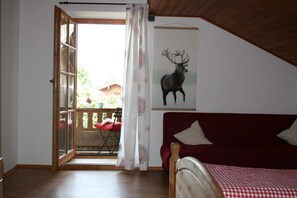 A bedroom with a wooden ceiling, a red sofa, a bed with a checkered bedspread, a large window with a view of the outdoors, and a deer painting on the wall.