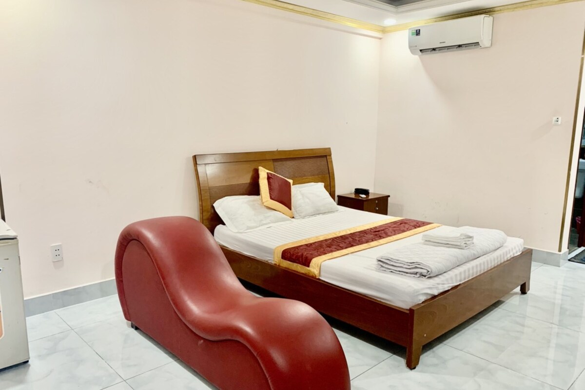 A hotel room with a bed, a red chair, a nightstand, and an air conditioning unit.