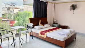 Una habitación de hotel con cama, una mesita, una silla, una mesita de noche y un ventilador de pared.