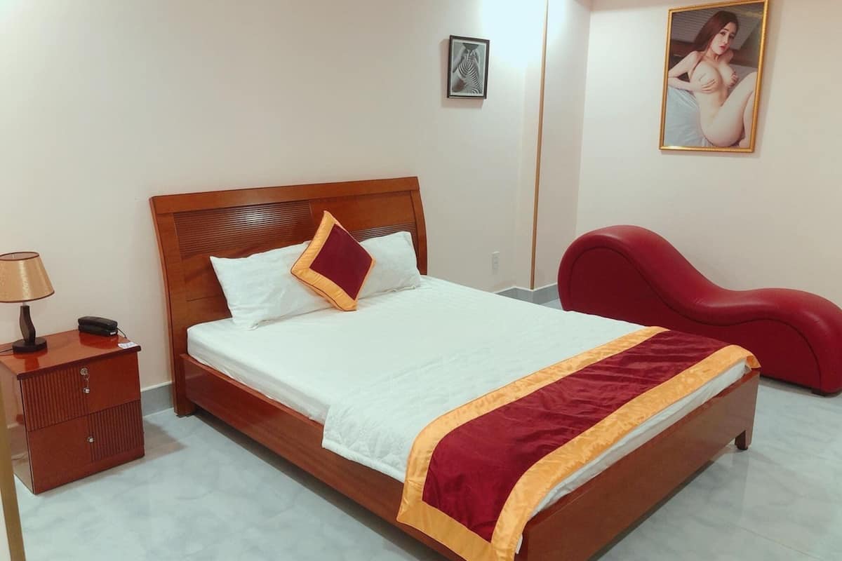 A neatly made bed with a wooden headboard, a red chair, a bedside table with a lamp, and a framed picture on the wall.