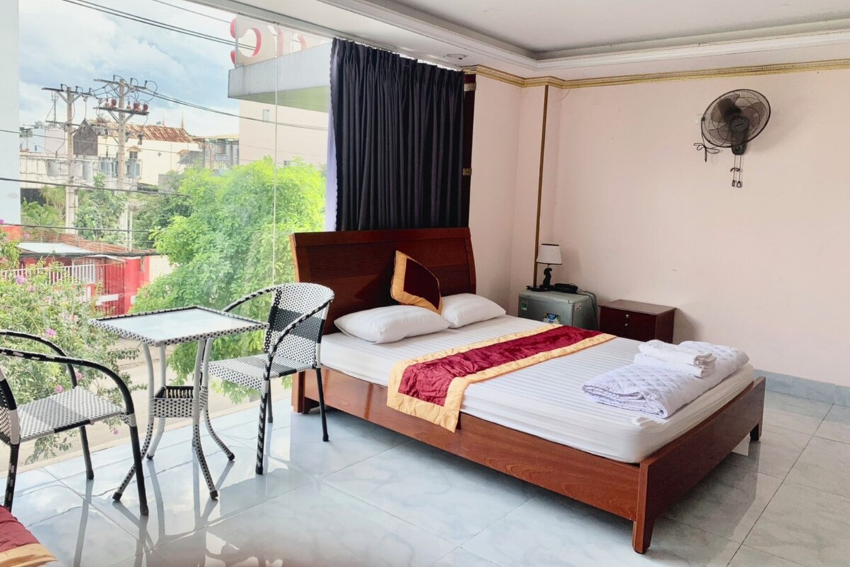 A hotel room with a bed, a small table, a chair, a nightstand, and a wall-mounted fan.