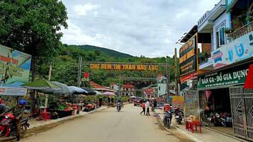 Um mercado de rua com diversos quiosques e lojas, um letreiro em destaque para 'CHO DEM THI TRAN BAO LAC', e um cenário montanhoso ao fundo.