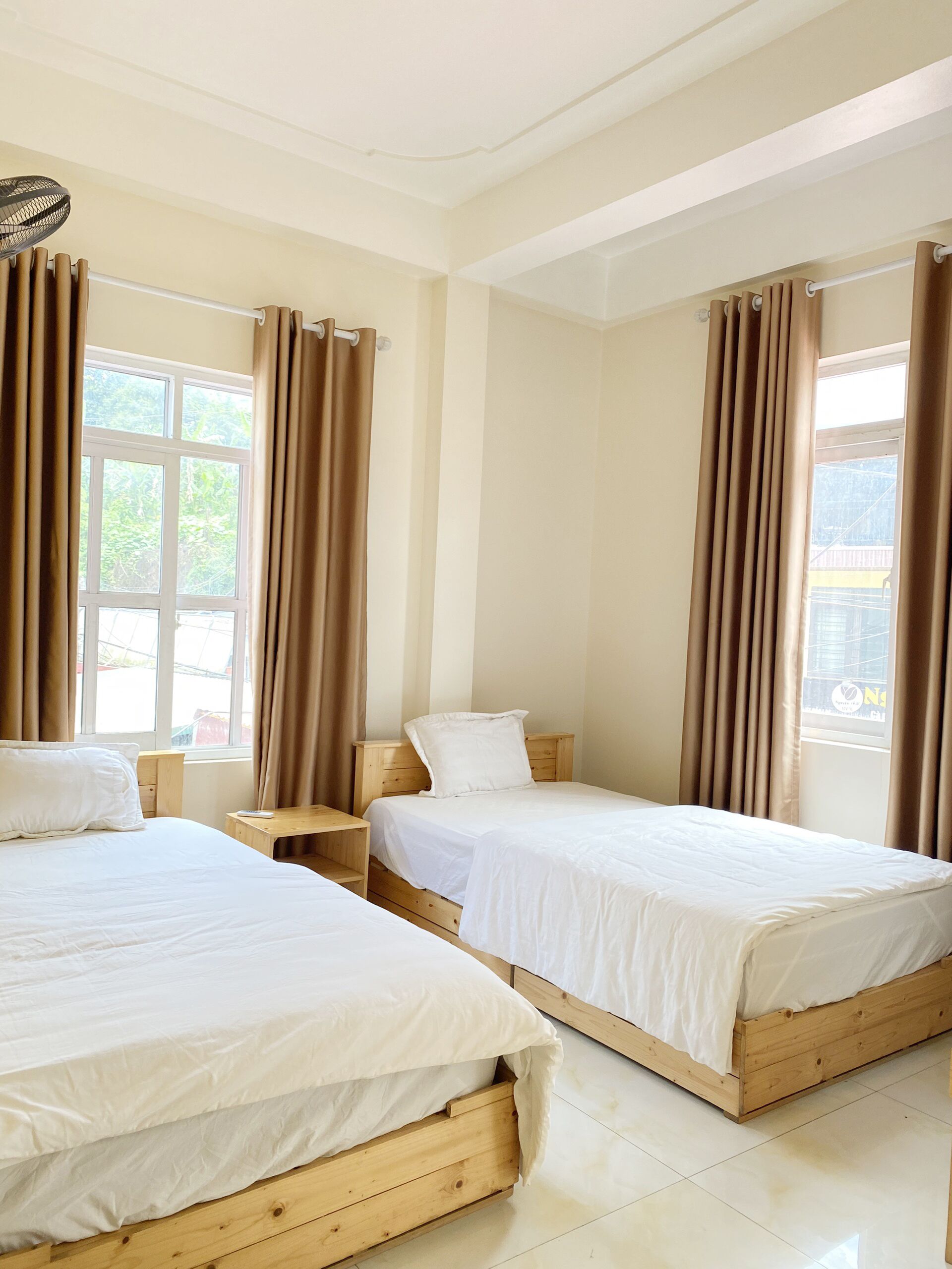 Una habitación de hotel con dos camas de madera, una mesita de noche de madera y un ventanal grande con cortinas.