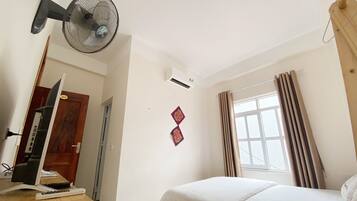 A bedroom with a bed, a desk with a computer, a standing fan, an air conditioning unit, and a window with curtains.