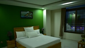Quarto de hotel com cama de madeira, roupa de cama branca, parede verde, um quadro, mesa de cabeceira com abajur e janela com cortinas.