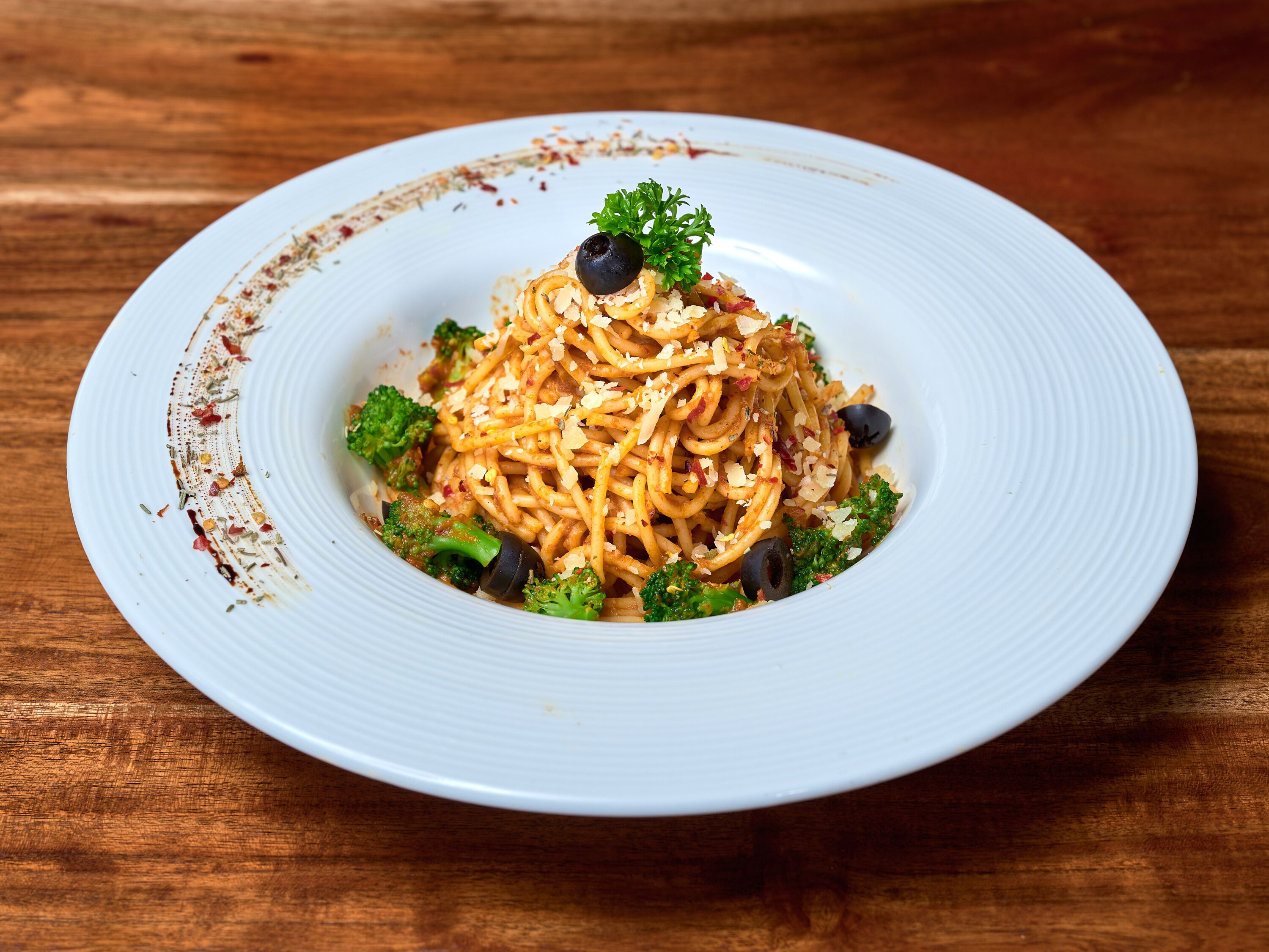 A white plate with spaghetti, broccoli, and black olives on a wooden table.