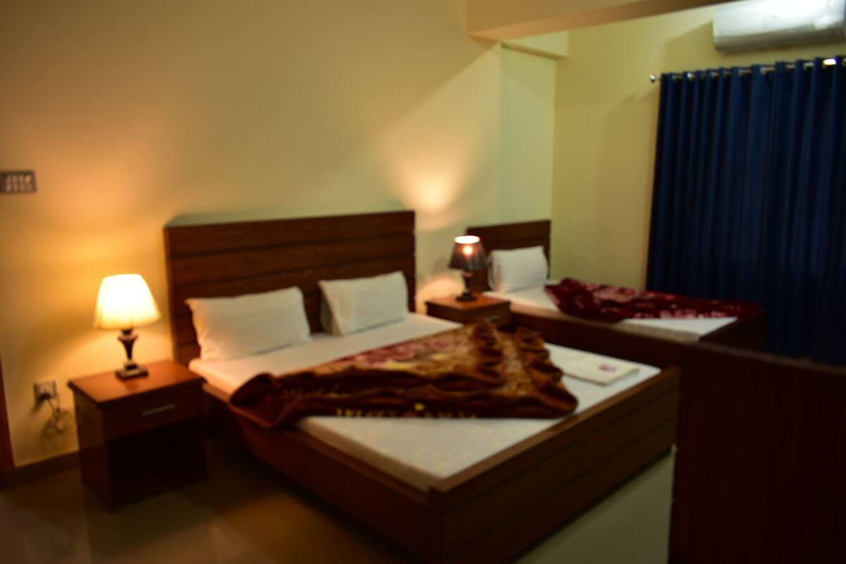 Une chambre d’hôtel avec un lit, une table de chevet avec une lampe, une commode et une fenêtre avec des rideaux.