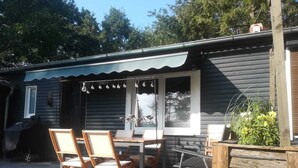 En træterrasse med et bord og stole, et vindue med gardiner, og et lille haveområde.