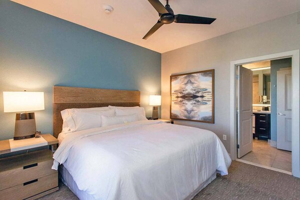 A hotel room with a bed, two bedside tables with lamps, a ceiling fan, and a painting on the wall.
