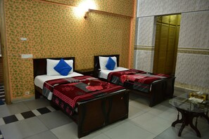 A room with two beds, each with a pillow and a blanket. There is a small table with a glass top and a chair. The walls have a floral wallpaper pattern. - State Continental Guest House (Mekhtar)
