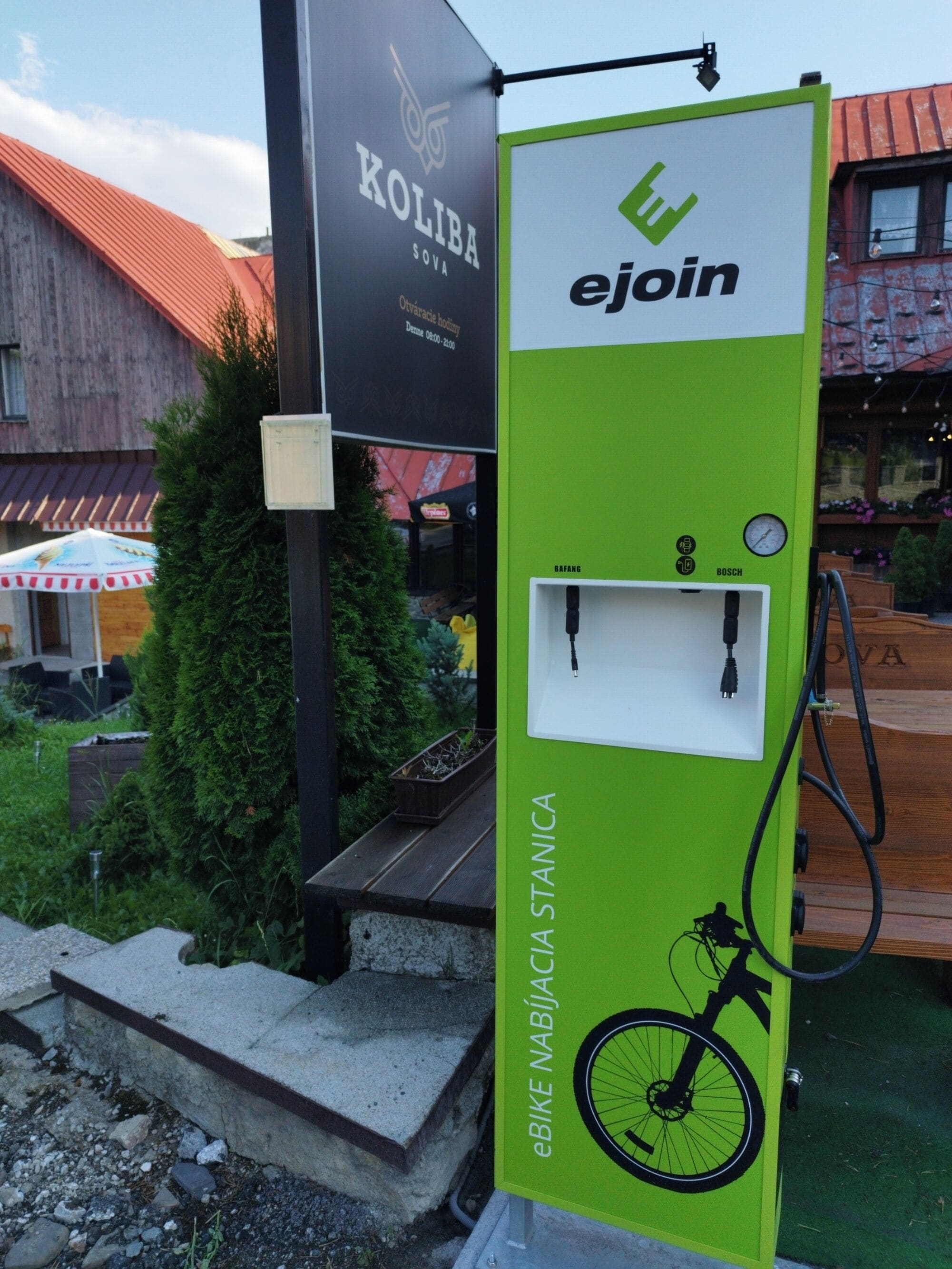 Une station de recharge pour vélos électriques, de couleur verte et blanche, située à l’extérieur, avec un bâtiment en bois et un arbre en arrière-plan.