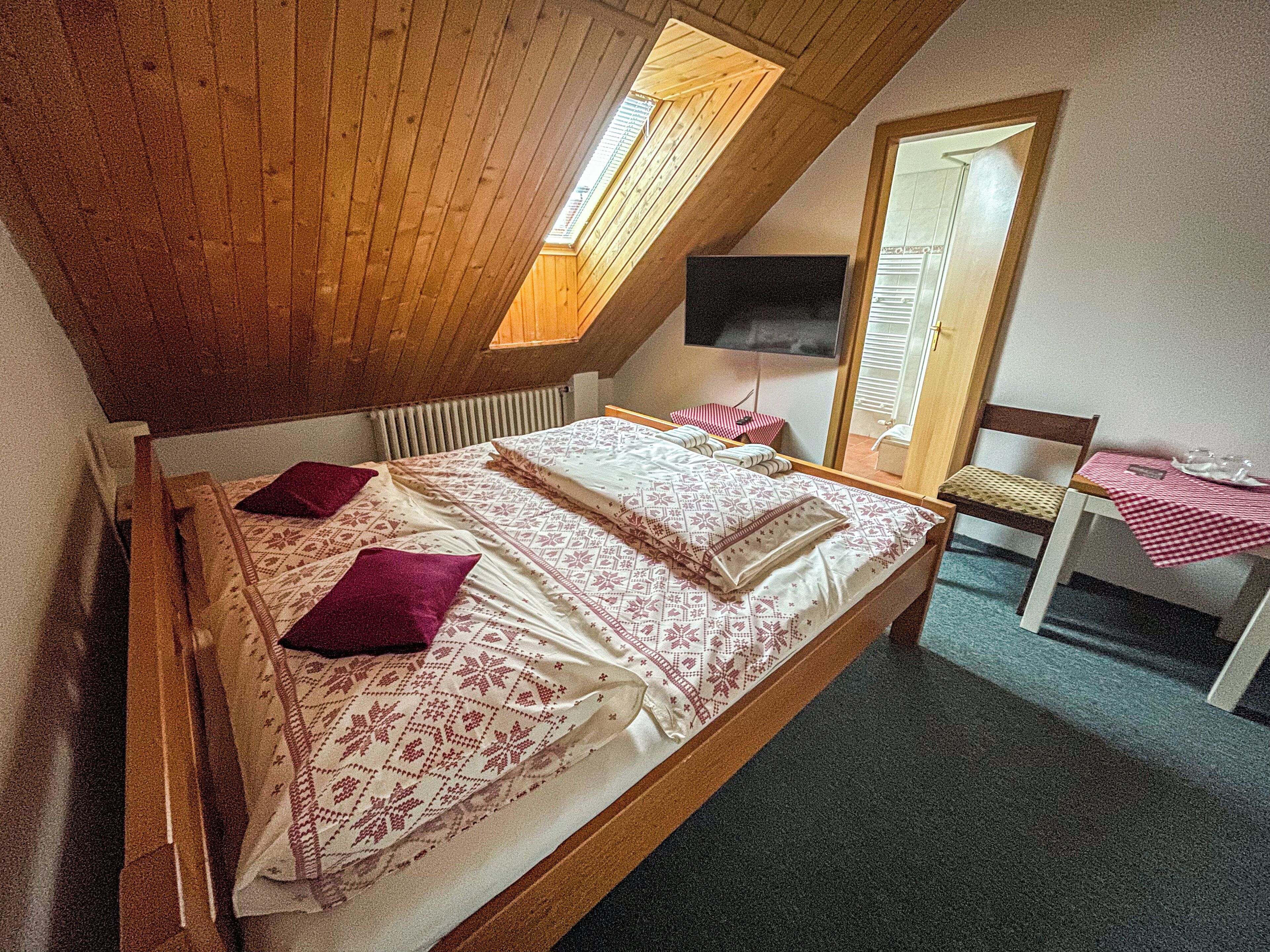 A bedroom with a sloped ceiling, a bed with patterned bedding, a chair, a small table with a checkered tablecloth, and a TV mounted on the wall.
