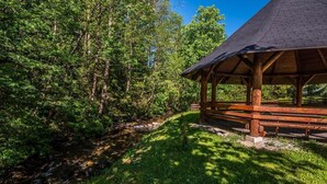 A wooden pavilion with a sloped roof, surrounded by lush greenery and a small stream. - Hotel Sova Ždiar (Zdiar)