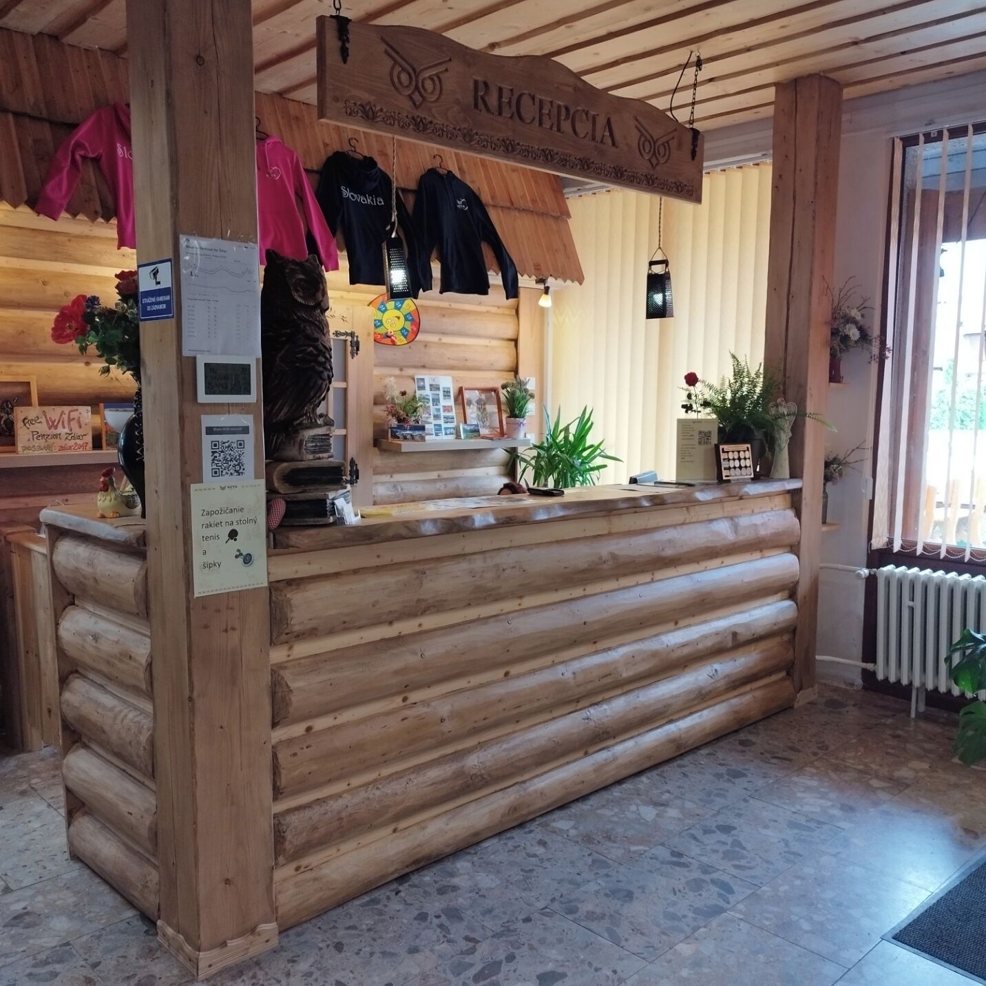 Um balcão de madeira na receção com um letreiro a dizer "RECEPCIA", um porta-casacos com blusas, uma pequena estátua e um código QR para acesso à rede WiFi.