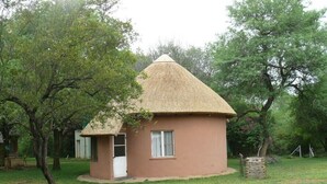 Une petite maison à un seul étage, avec un toit de chaume, entourée d’arbres et d’herbe.