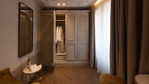 Une chambre d’hôtel avec un plancher en bois, un miroir, une petite table sur laquelle se trouvent une bouteille et une carte, une armoire à portes à persiennes et une grande fenêtre avec des rideaux.