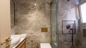 A modern bathroom with a marble wall, a glass shower enclosure, a white sink, and a gold-toned vanity.