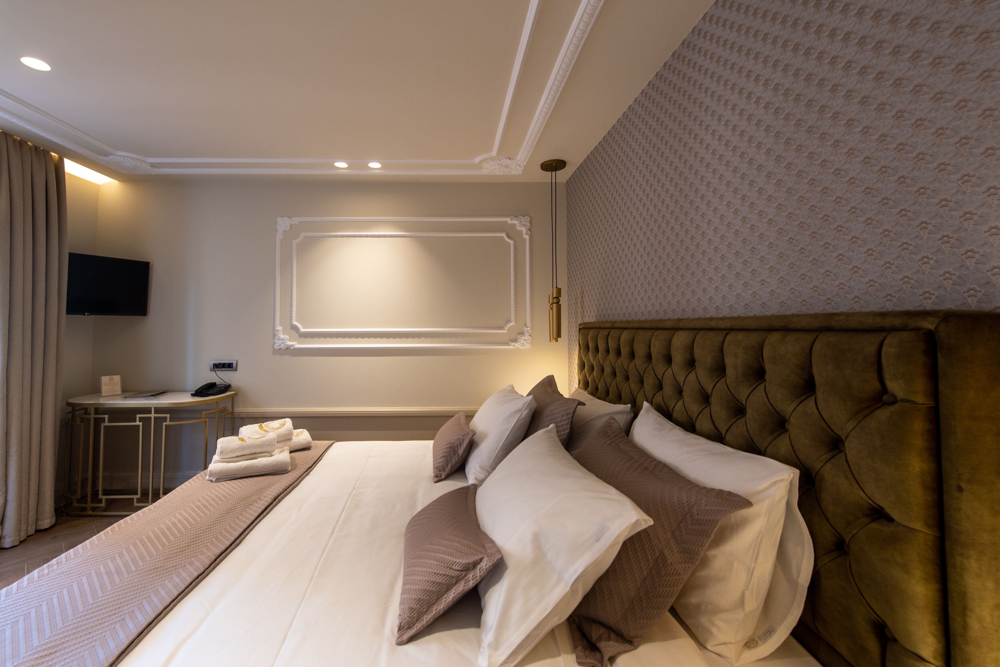 Una habitación de hotel con una cama grande, un televisor de pantalla plana montado en la pared, una mesita de noche con lámpara y un papel tapiz estampado.