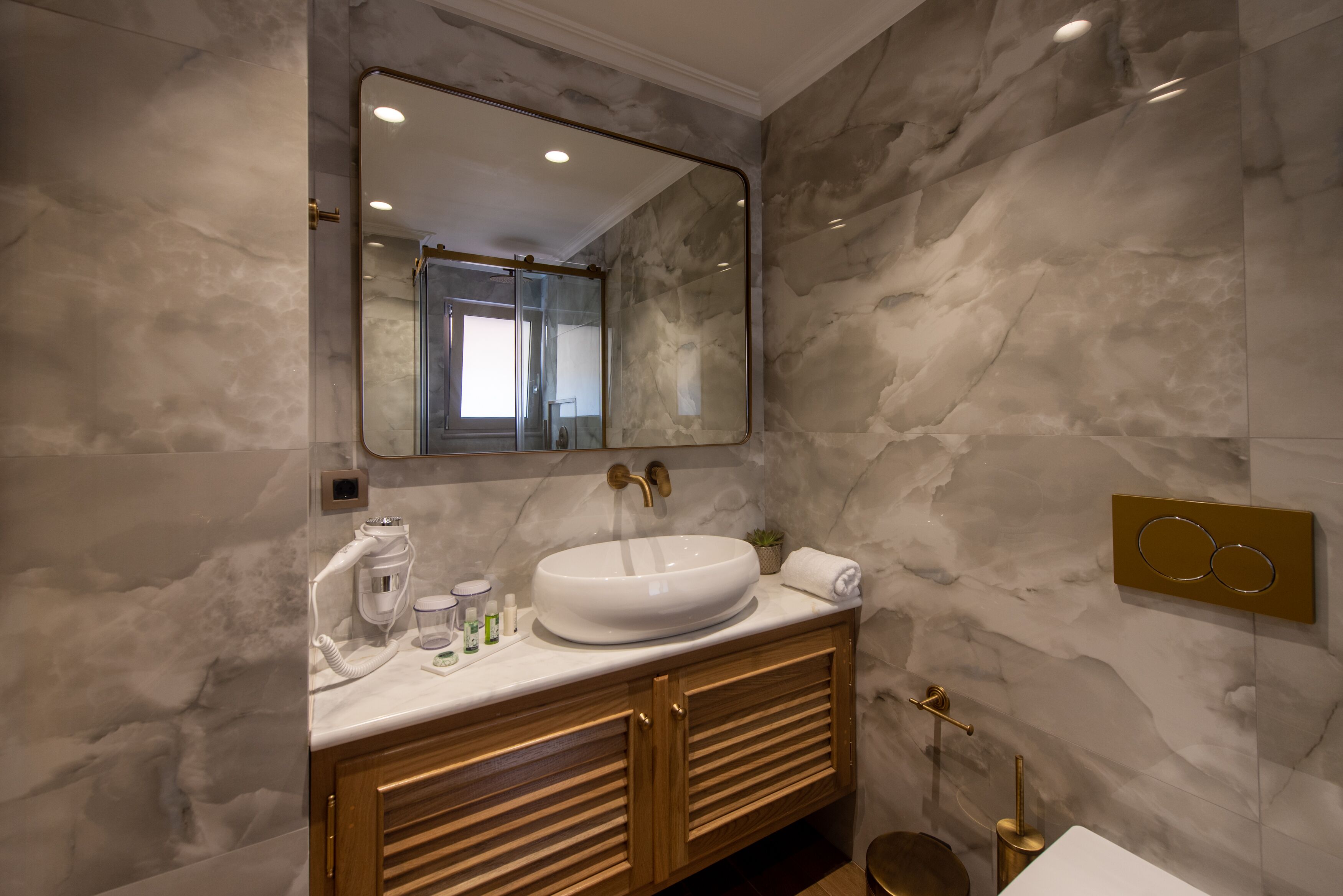 Een moderne badkamer met een marmeren wand, een grote spiegel, een wit wastafelbakje, houten kastjes en een gouden kraan.