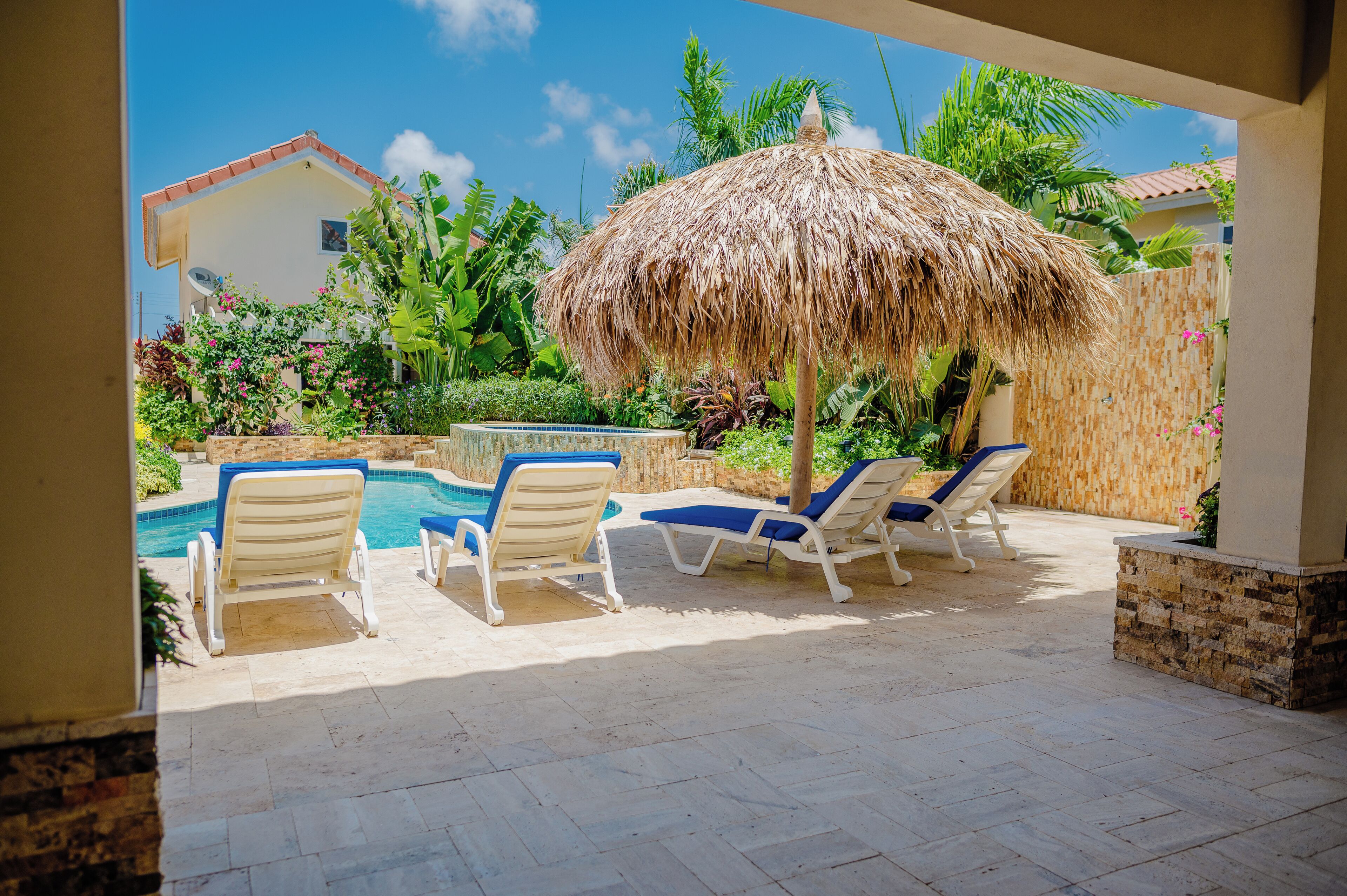 A poolside area with lounge chairs and a thatched umbrella, surrounded by lush greenery and a house in the background.