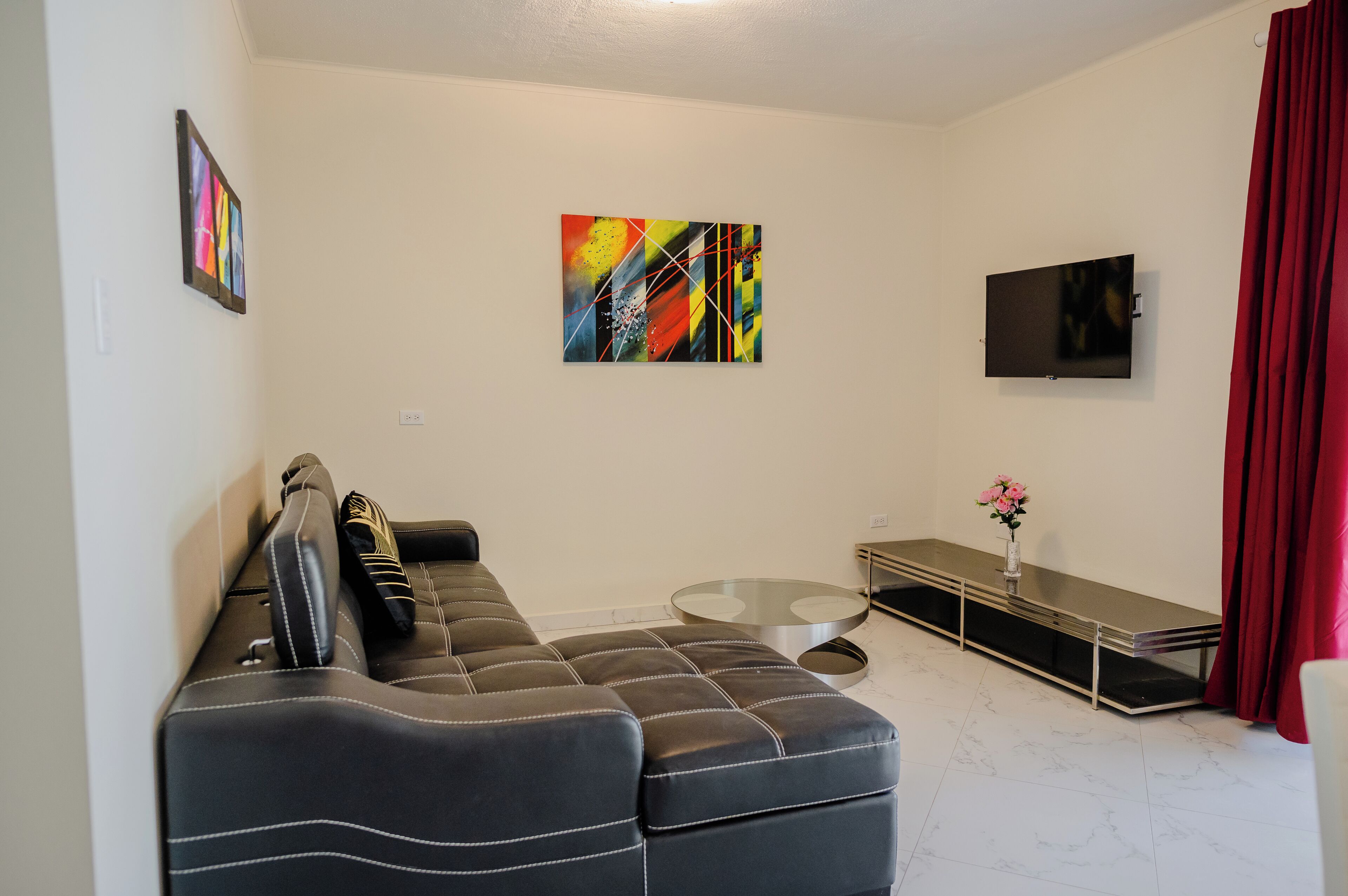 Ein modernes Wohnzimmer mit einem schwarzen Ledersofa, einem Glastisch, einem Flachbildfernseher und einem roten Vorhang.