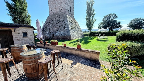 Un espace patio agrémenté de barils en bois, d’une table et de chaises. On aperçoit un mur de pierre et une grande tour en arrière-plan.