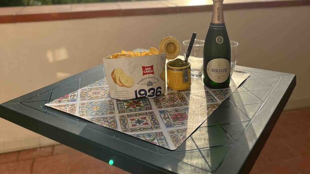 A table with a bottle of Berlucchi wine, a jar of olives, and a box of San Carlo chips on a balcony with a scenic mountain view in the background. - A Refuge Between Green And Blue (Tramonti)