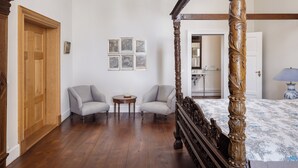 Ein Schlafzimmer mit einem Himmelbett, zwei Sesseln, einem kleinen runden Tisch und einer Holztür.