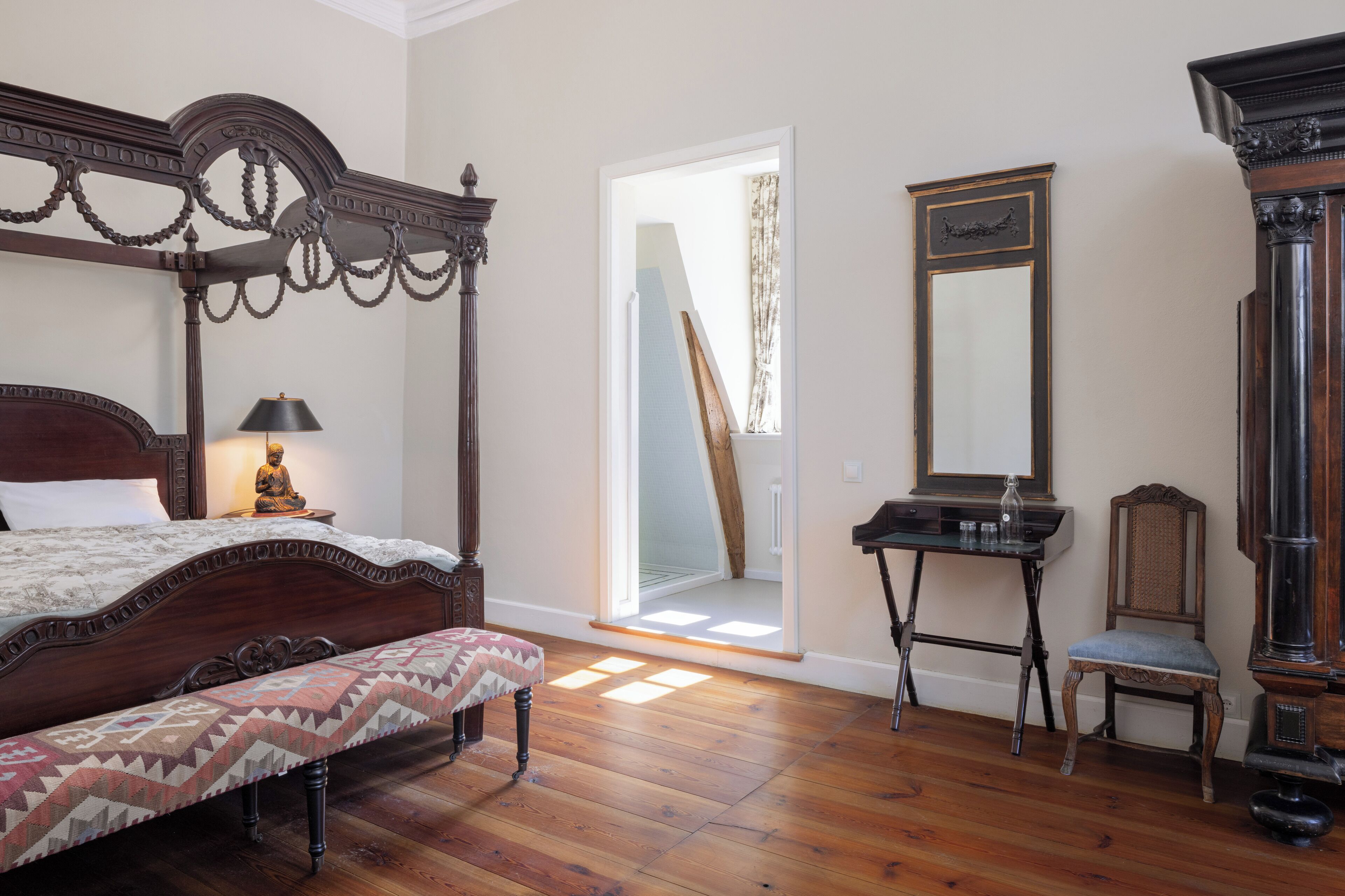 A bedroom with a wooden bed, a bench, a desk, a chair, and a large mirror.