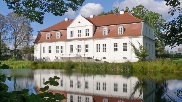 Ein großes, weißes Gebäude mit rotem Dach, das sich in ruhigem Wasser spiegelt.