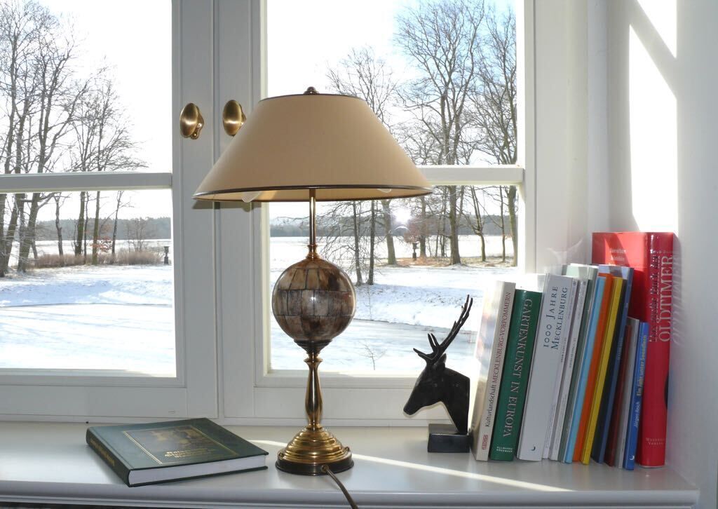 Een bureau met een globe lamp, boeken en een hertenornament bij een raam met uitzicht op een besneeerd landschap.