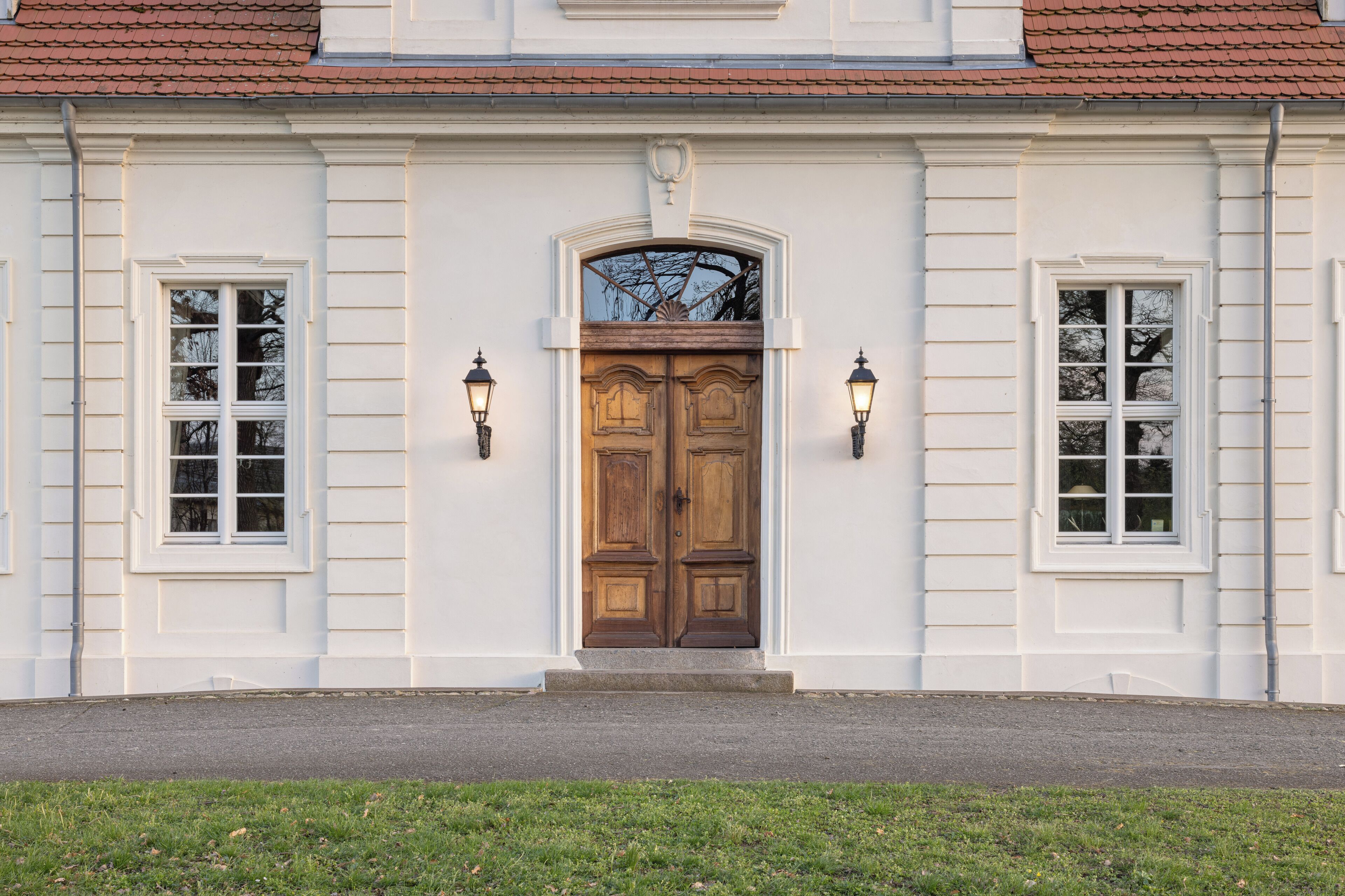 A building with a central wooden door, flanked by two windows with white frames and two wall-mounted lamps.