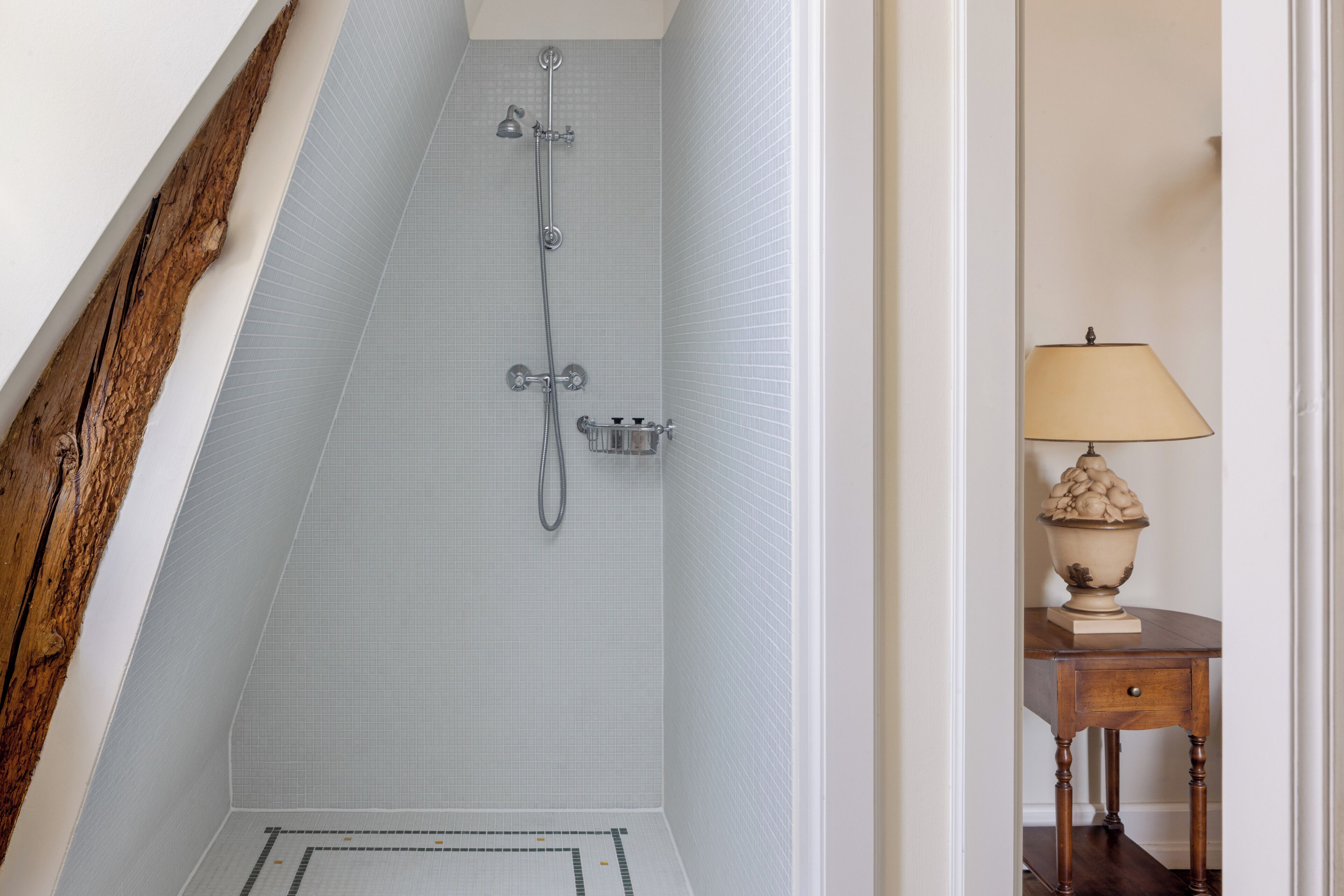 Un baño estrecho con pared de azulejos, una viga de madera y una pequeña mesa con lámpara.