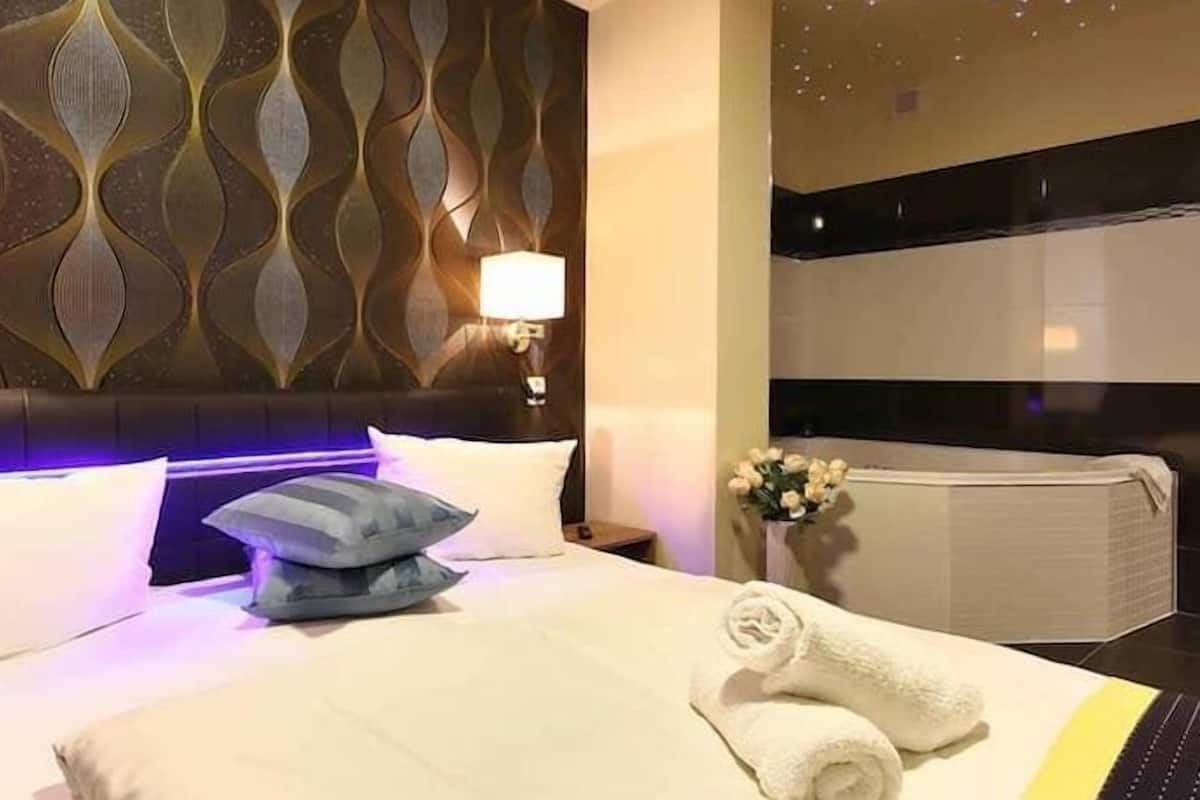 A hotel room with a modern design, featuring a patterned headboard, a neatly made bed with white and gray bedding, and a bedside table with a lamp.