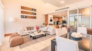 A modern living room with a sofa, coffee table, and ottoman, a dining table with chairs, and a kitchen in the background. - Luxury Golf View Apartment near Casa de Campo (Casa De Campo)