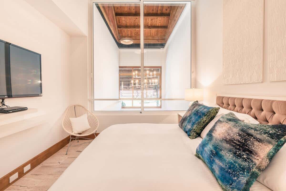 Ein Schlafzimmer mit einem großen Bett, einem Fernseher auf einem Regal, einem Sessel und einem Fenster mit Blick auf ein Gebäude.