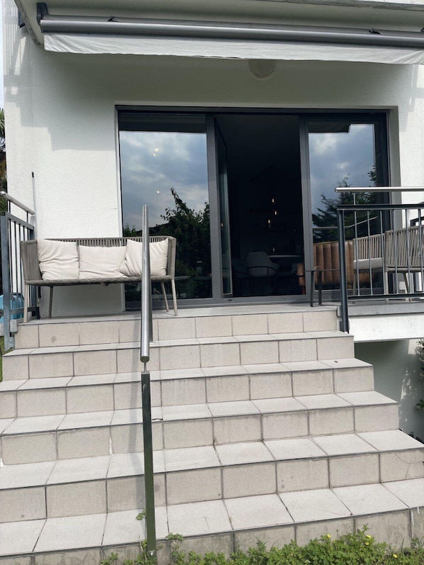 A modern building with a set of outdoor stairs leading to a glass door entrance, a white outdoor sofa, and a metal railing.