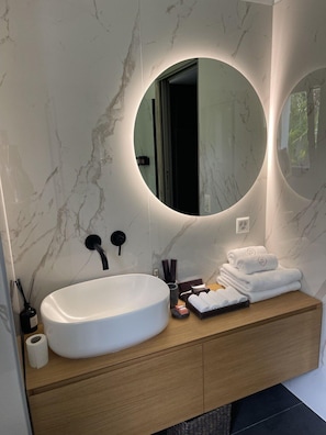 A modern bathroom with a marble wall, a large oval sink, a wooden vanity, and two round mirrors with under-cabinet lighting.