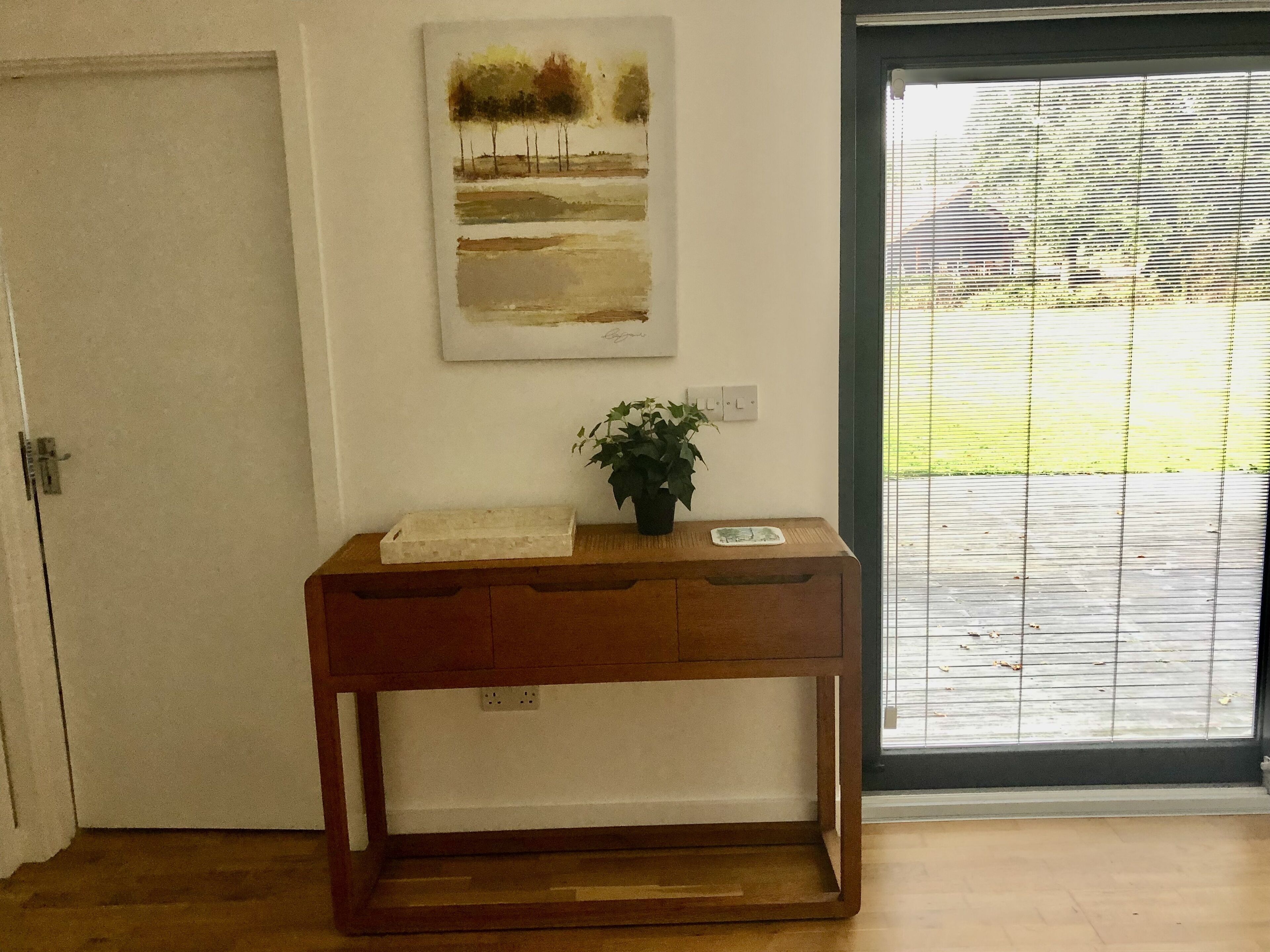 Una consola de madera con una planta y una bandeja pequeña, ubicada frente a una puerta con un cuadro en la pared.