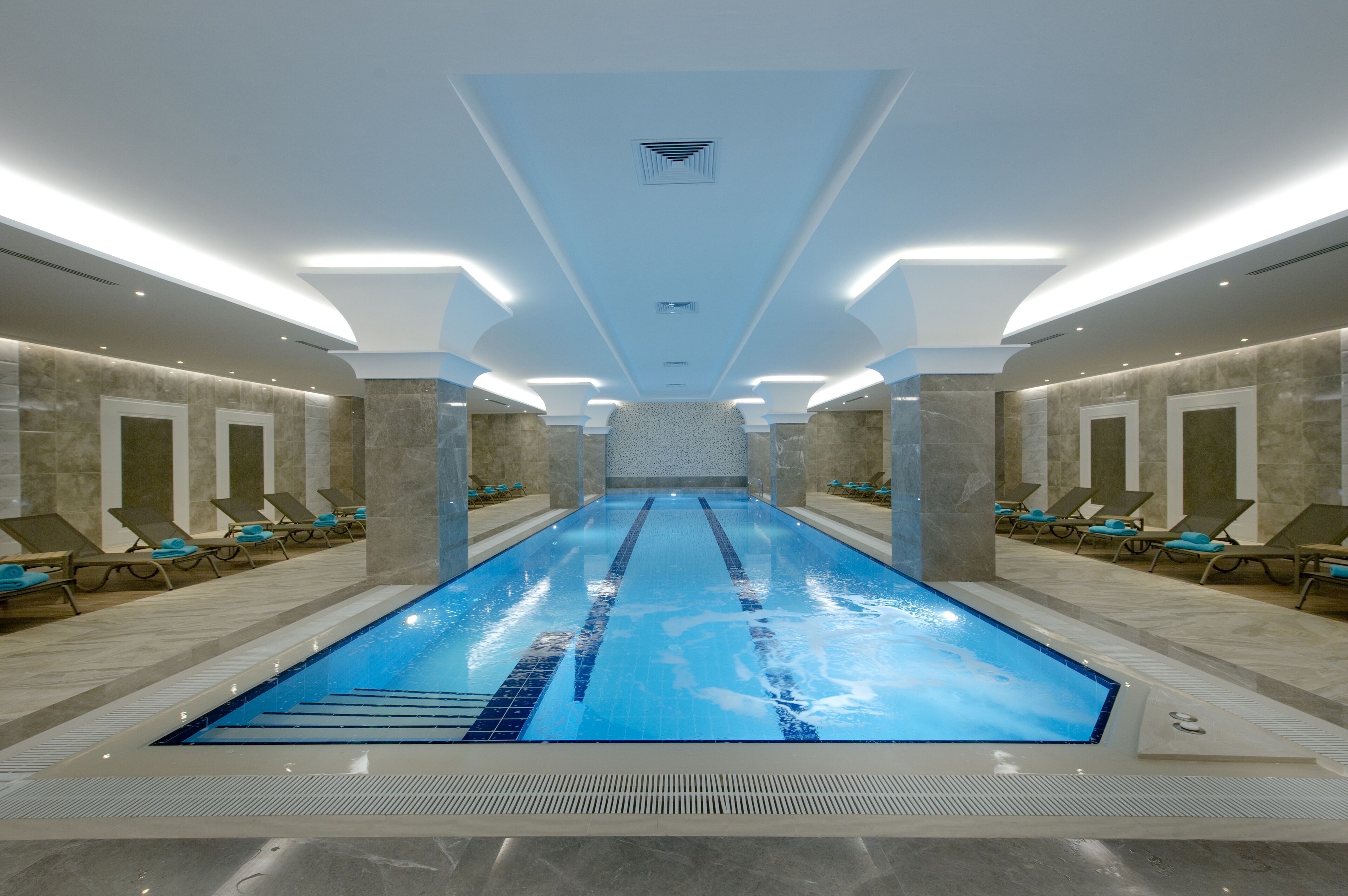 A long indoor swimming pool with a clear blue water, surrounded by lounge chairs and a modern ceiling design.