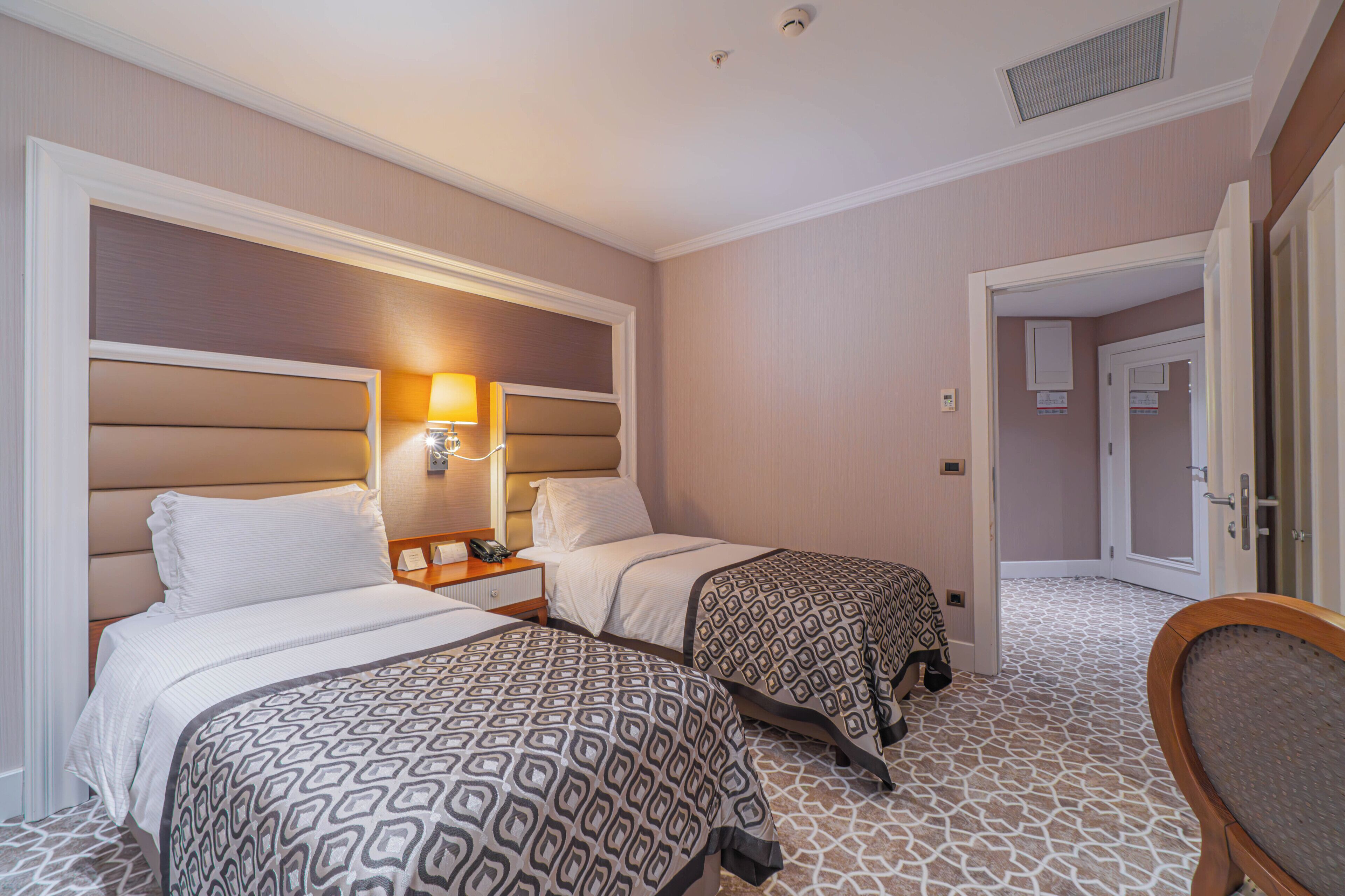 Ein Hotelzimmer mit zwei Betten, einem Nachttisch, einer Lampe und einer Tür zu einem weiteren Raum.