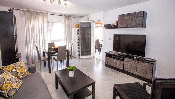 A modern living room with a sofa, a coffee table, a TV, and a dining area with chairs and a table. - Alcazaba Square Central Beach (La Herradura)