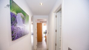 A hallway with a painting of a chair and flowers on the wall, leading to a room with a wooden door and a light switch on the wall. - Alcazaba Square Central Beach (La Herradura)