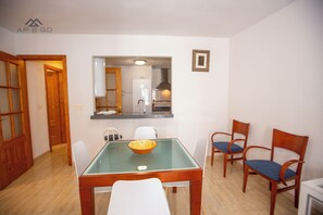 A dining area with a glass-top table, white chairs, and a wooden chair with a blue cushion. A wooden door and a white refrigerator are visible in the background.