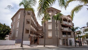 Un immeuble à plusieurs étages doté de balcons, de palmiers et d’une entrée sécurisée par un portail.