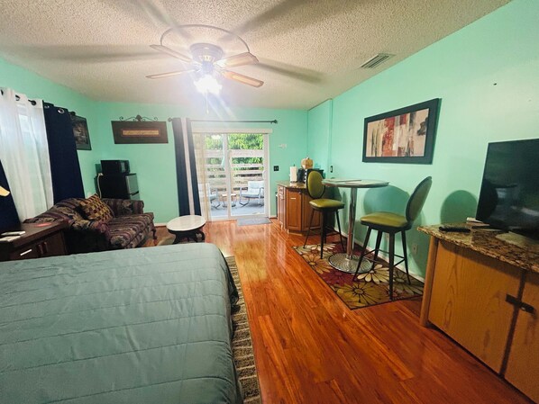 Ein kleines Zimmer mit einem Bett, einem kleinen Tisch, einem Sessel, einem Fernseher und einem Deckenventilator.