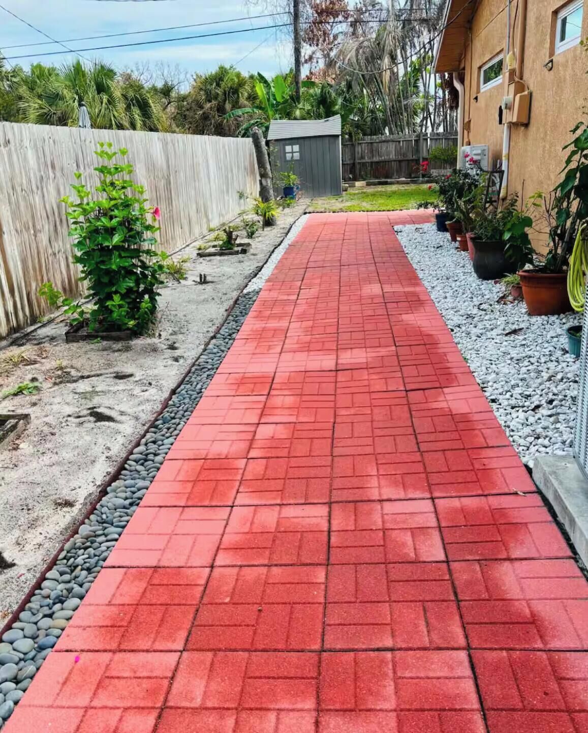 A red brick pathway leading to a house with a wooden fence and potted plants on the side.