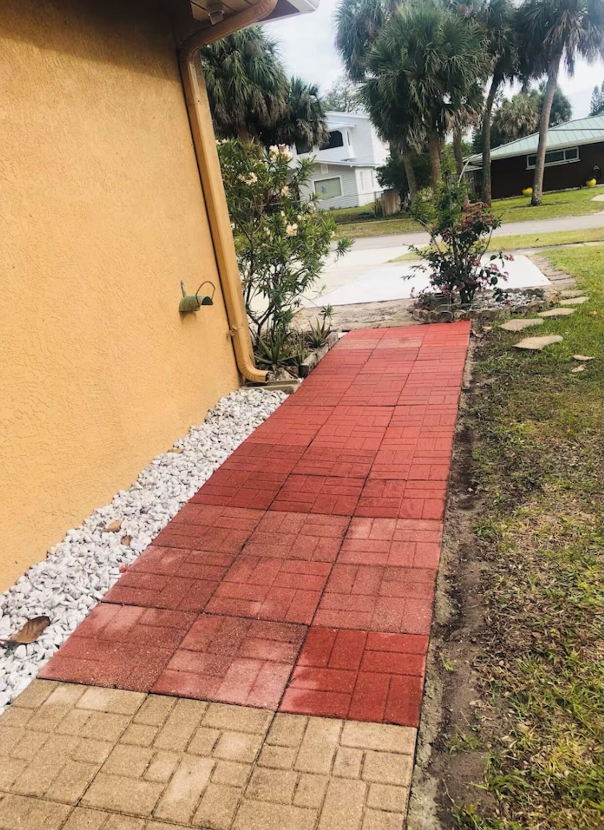 A red brick pathway leading to a building with a yellow wall and a small garden area with various plants.